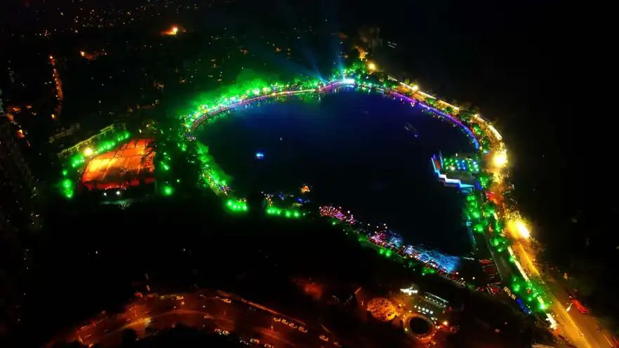 Night aerial view of a lakeside festival, the shore ringed with green and violet lights