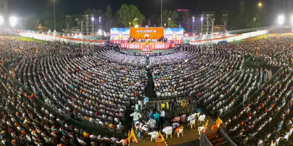 A floodlit rally ground packed with seated crowds facing a large stage with LED screens