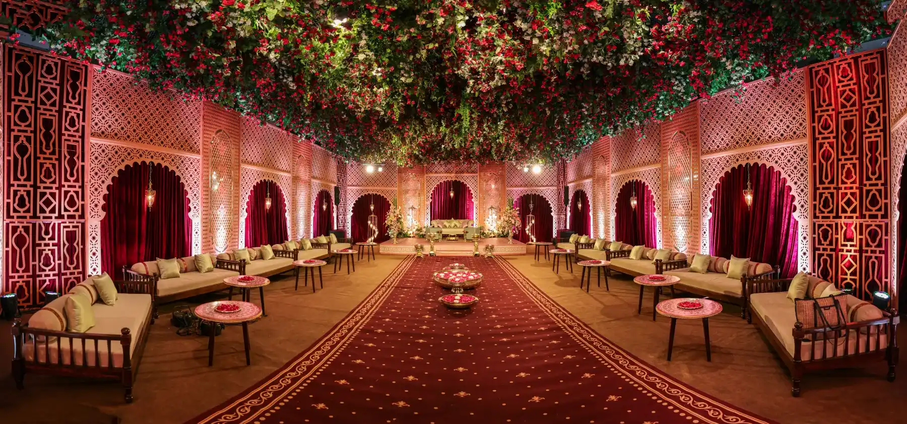 An ornate indoor wedding mandap with a floral ceiling, lounges and a red carpet